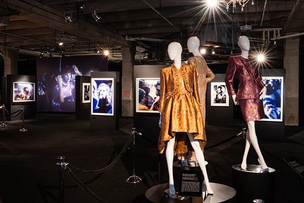 Dark gallery with three brightly lit couture ensembles on mannequins, photographs