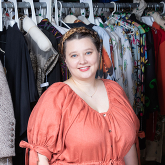 Ailie in front of a rack of hanging garments, smiling and wearing an orange top with her hair up